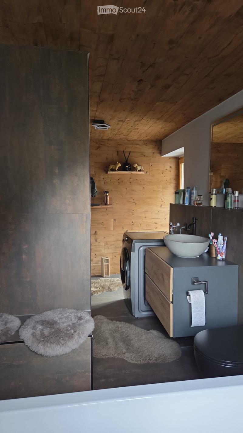 Modern bathroom with wooden walls, a large bathtub, a sink with a modern faucet, a toilet, and shelves holding toiletries. The floor is covered with plush, furry rugs, and the space features a sleek, contemporary design.