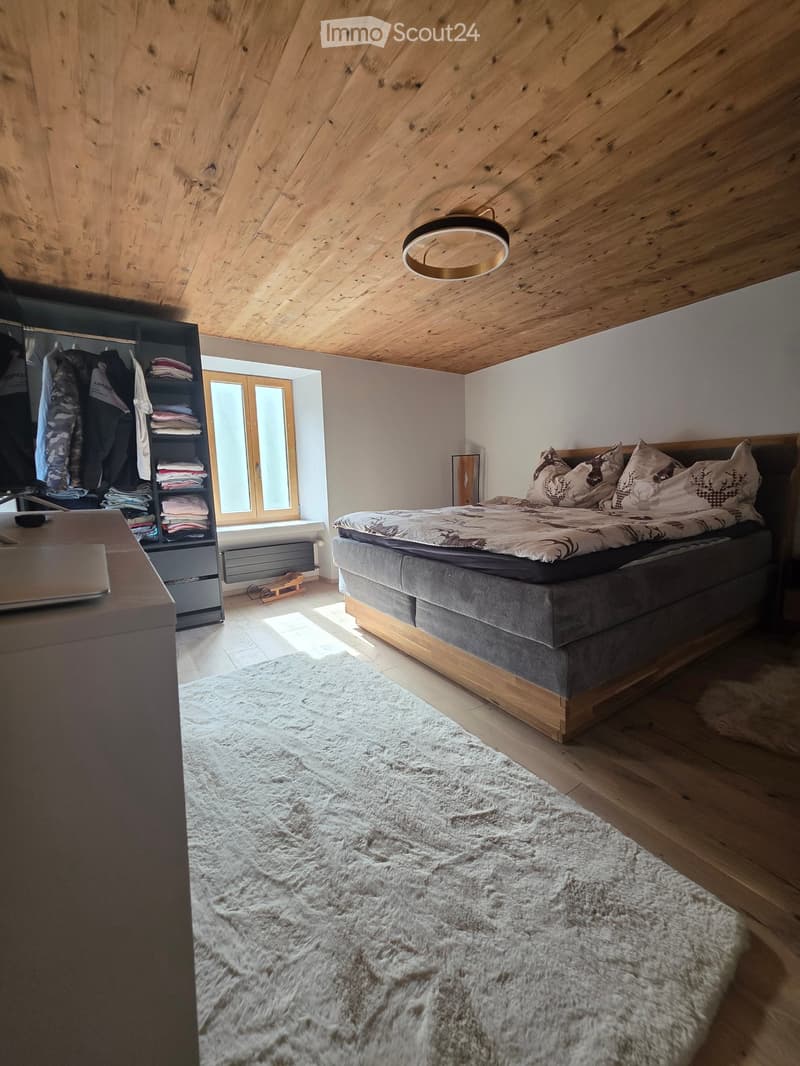 Cozy bedroom with wooden slanted ceiling, large window, bed with patterned bedding, white shag rug, wardrobe, and laptop on a desk.