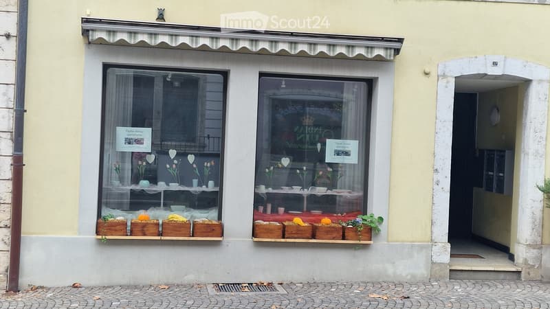 Facade of a building with two large windows displaying ceramic items and floral arrangements, with a striped awning above.
