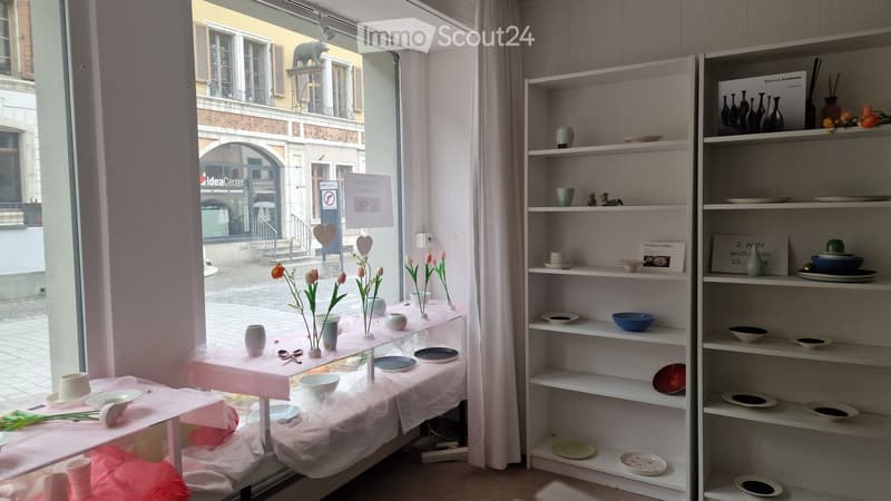 A well-lit room with white walls and large windows, featuring shelves filled with ceramic plates, bowls, and vases, and a small table with flowers and ceramic items.