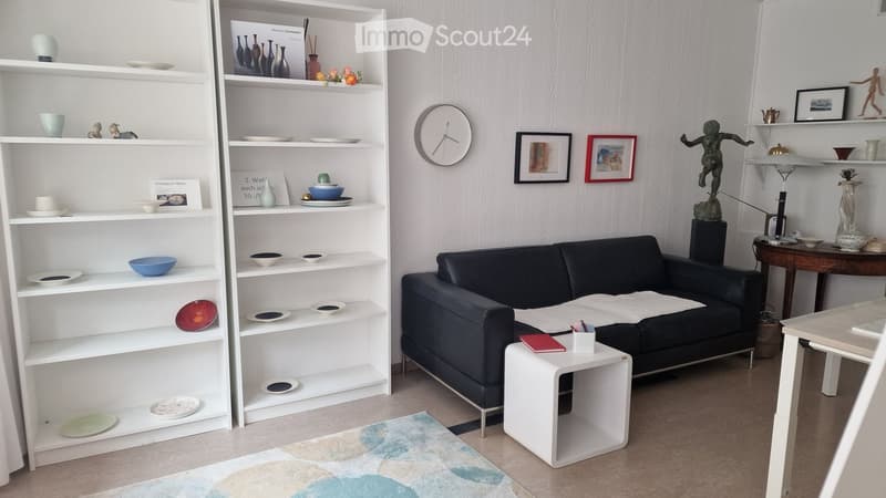 Modern living room with a black leather sofa, white coffee table, white shelving unit with decorative items, a clock on the wall, and a small table with a lamp.