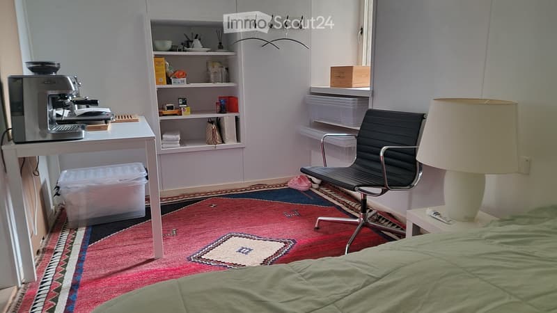 A small, furnished room with a bed, a desk with a coffee maker, a chair, a lamp, and shelves holding various items.