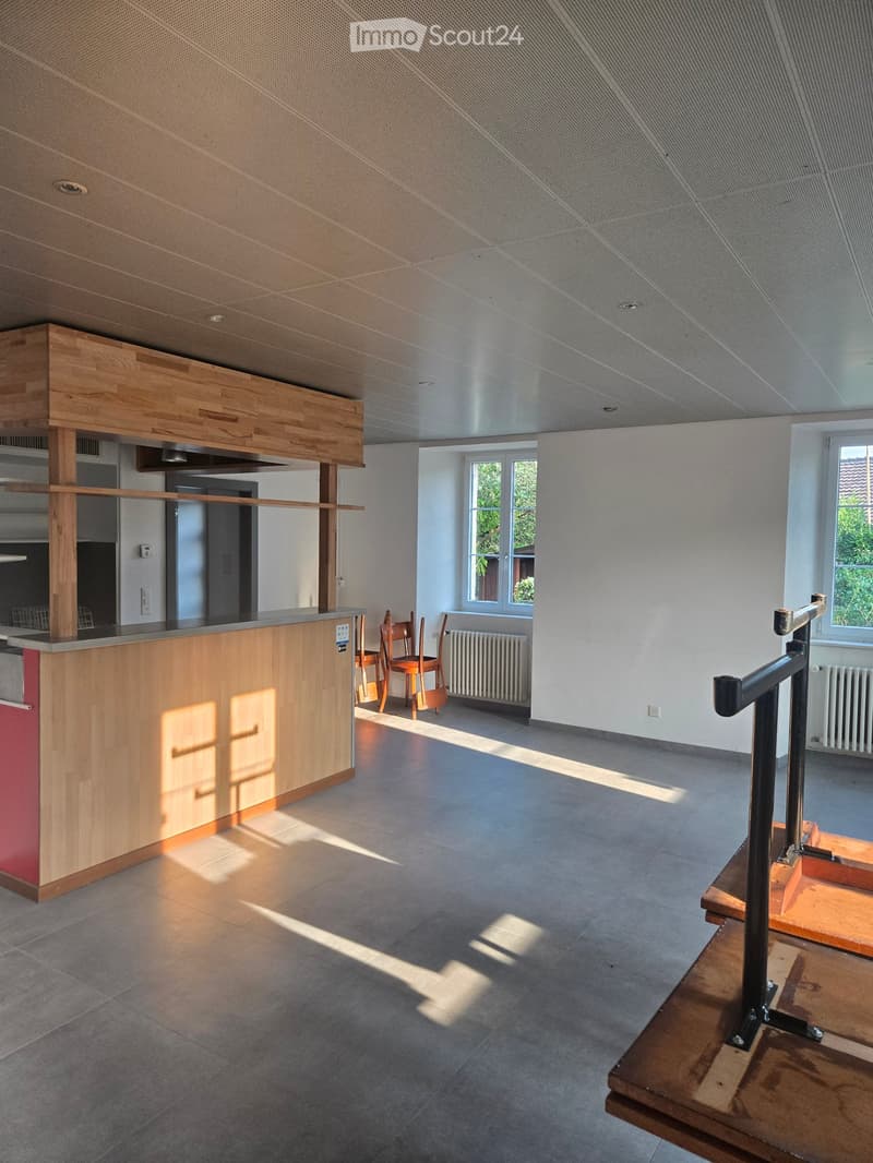Modern, open-plan room with wooden staircase, a small wooden table, and two chairs near a window. The space features a polished concrete floor and white walls.
