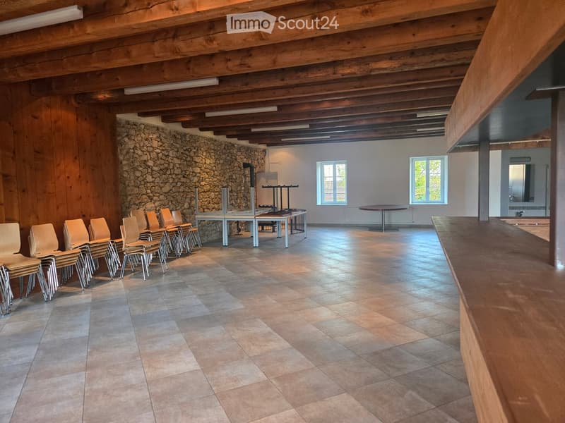 Empty room with wooden beams, stone wall, tiled floor, and a row of chairs and a table.