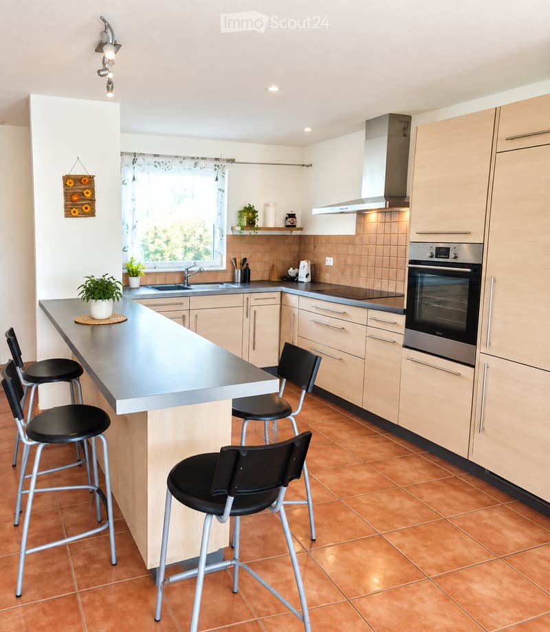 Modern kitchen with light wood cabinets, stainless steel appliances, a central island with bar stools, and a window overlooking greenery.