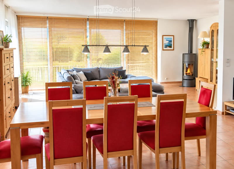 Bright living and dining area with a grey sofa, wooden dining table with red chairs, fireplace, and large windows with blinds.