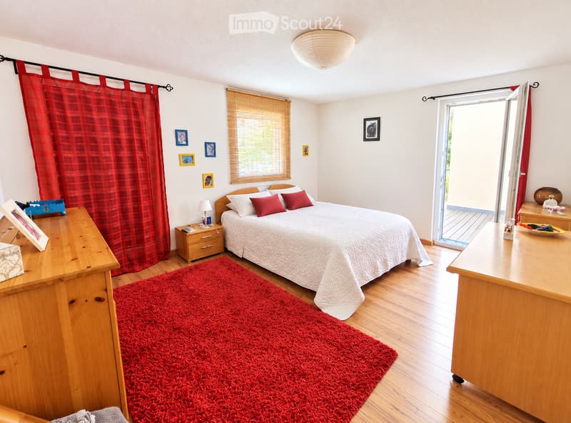 Cozy bedroom with wooden flooring, a large bed with white and red bedding, a wooden nightstand with a lamp, a wooden dresser, picture frames on the wall, red plaid curtains, and a red shag rug.