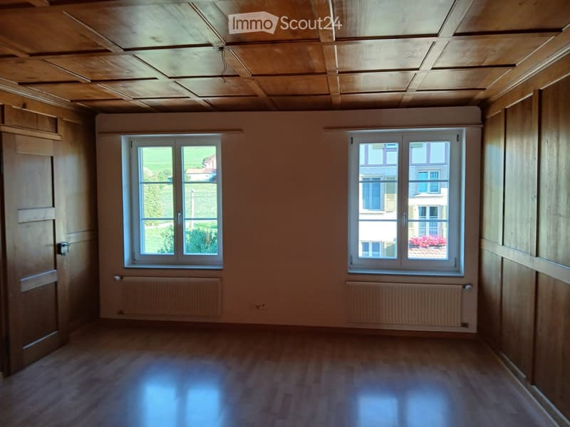Empty room with wooden flooring, wooden ceiling, two windows, radiators, and a door.