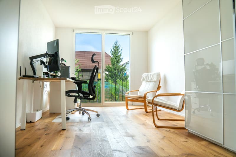 A modern, well-lit room with wooden flooring, a large window offering a view of greenery, a desk with a computer setup, and two comfortable chairs.