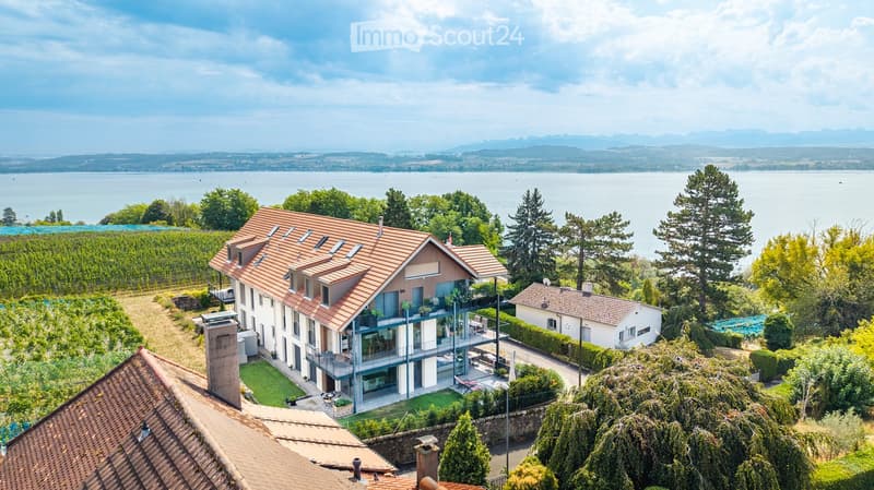 A modern two-story house with a red-tiled roof, large windows, and a spacious balcony overlooking a scenic lake and vineyard area.