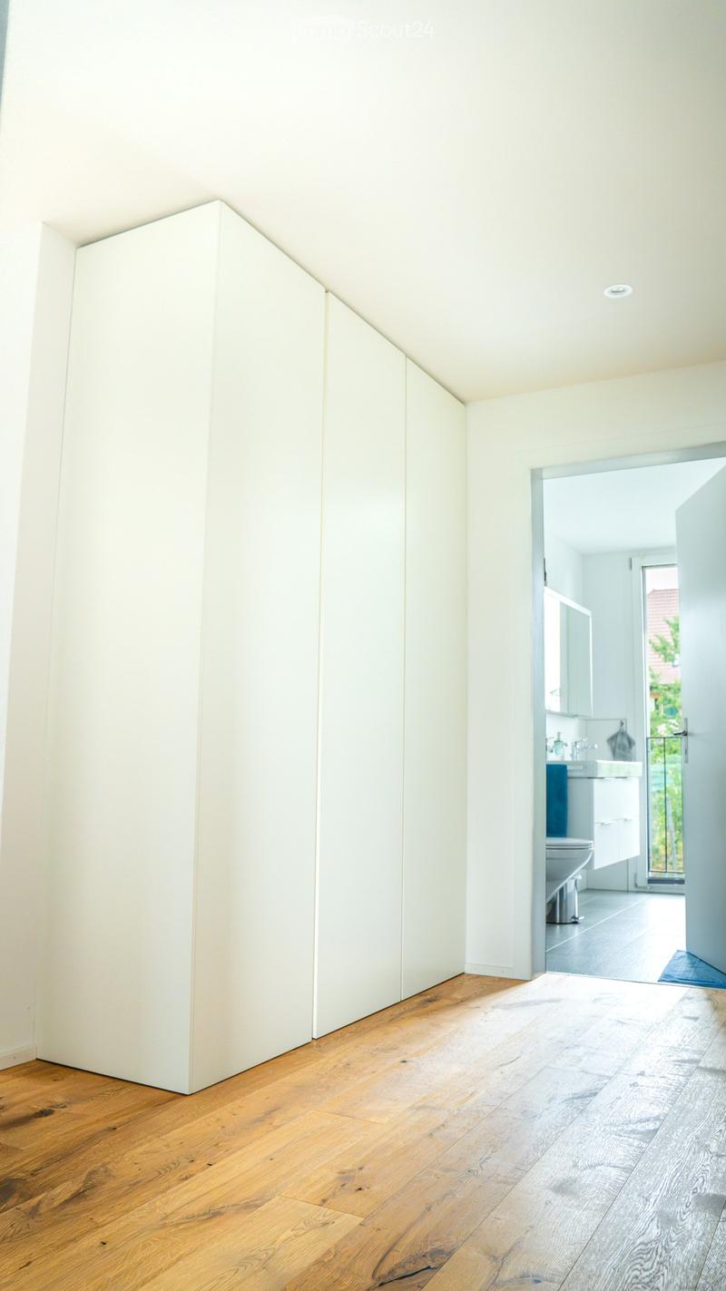 Bright hallway with wooden flooring, white walls, and a partially open door leading to a modern bathroom with white fixtures and a blue accent wall.