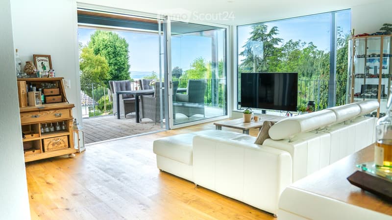 Modern living room with white leather sofa, coffee table, large TV, bar cabinet, and outdoor dining area visible through large glass doors.