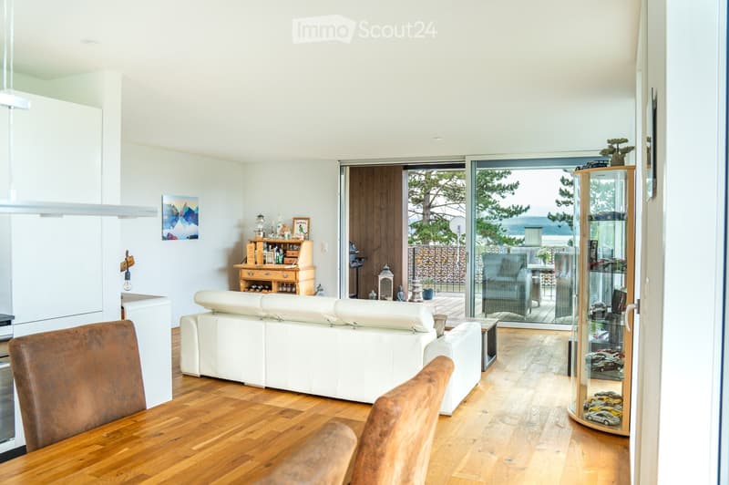 Bright living room with white walls, wooden flooring, and large sliding glass doors leading to a balcony. Features a white leather sofa, wooden dining table with chairs, a wooden cabinet, and decorative items.