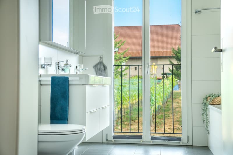 Modern bathroom with white tiles, a toilet, sink, and mirror. A large window offers a view of a vineyard outside.