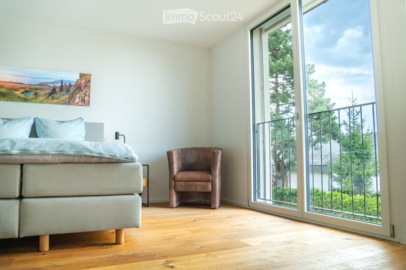 Bright bedroom with wooden flooring, a large bed with light bedding, a brown armchair, a nightstand with a lamp, and a scenic landscape painting on the wall. Large glass doors open to a balcony with a view of trees and a clear sky.