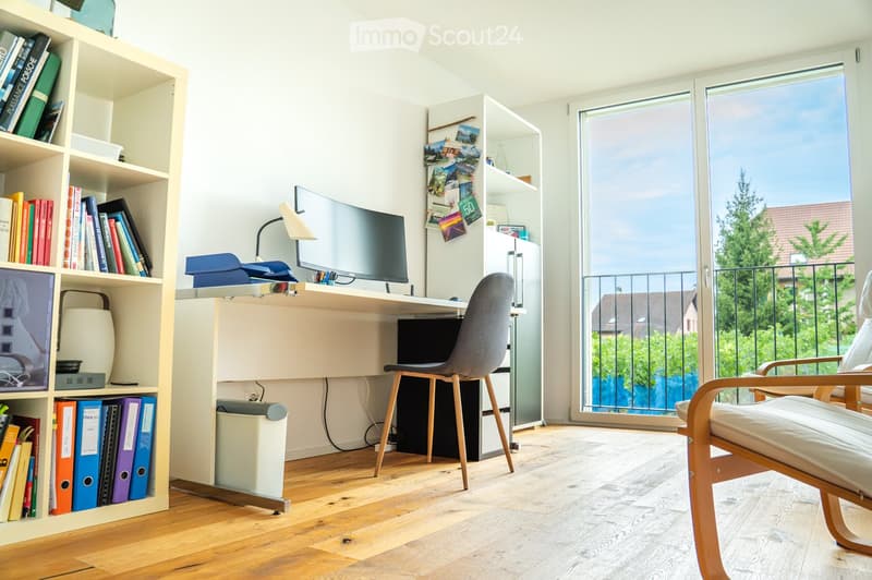 Bright, modern room with a desk, chair, screen, lamp, bookshelf, and a comfortable chair near a large window offering a view of the outdoors.