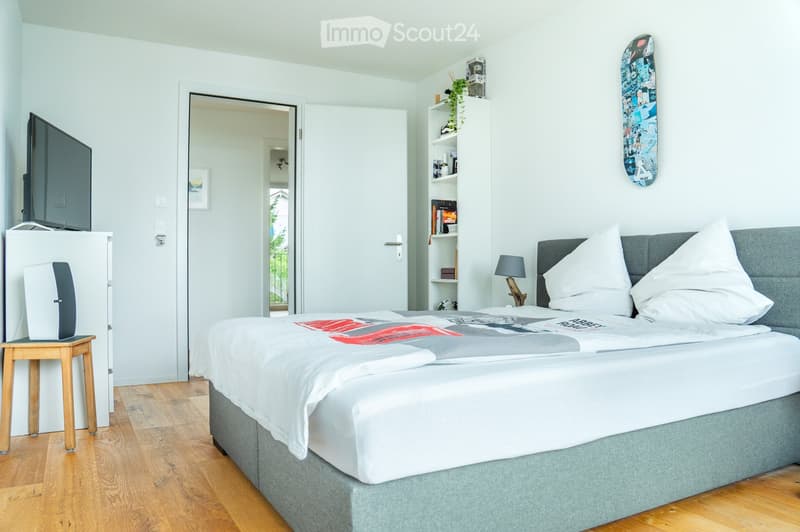 Modern bedroom with a large bed, nightstand, lamp, shelf, skateboard on the wall, and a screen on a small table.