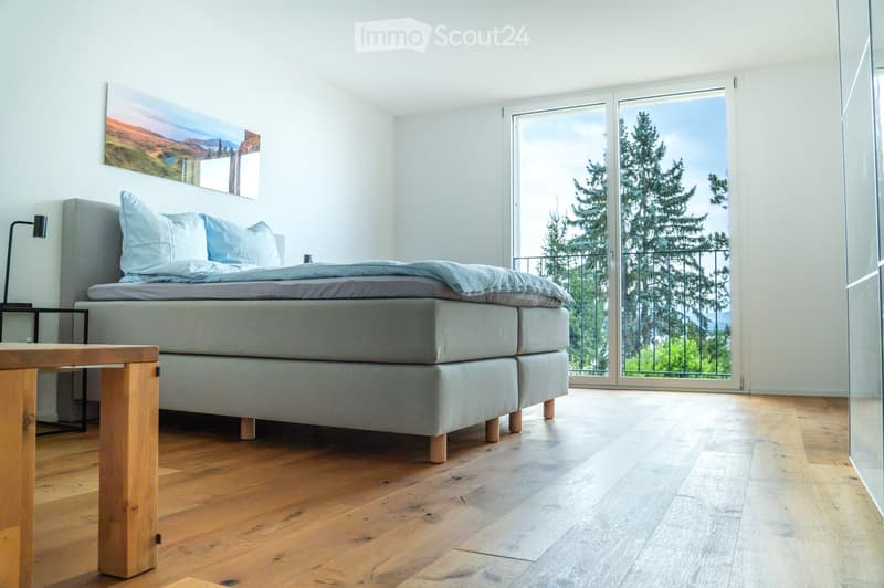 Modern bedroom with a large bed, nightstand, lamp, and a scenic view through large windows.