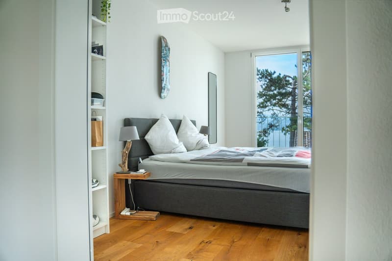 Modern bedroom with a grey upholstered bed, white pillows, wooden nightstand, and a view of trees through large windows.