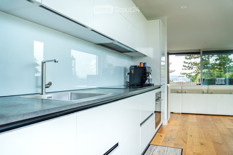 Modern kitchen with white cabinets, stainless steel sink, and a coffee machine on the counter. The space features a sleek design with a large window offering a view of the outdoor area.