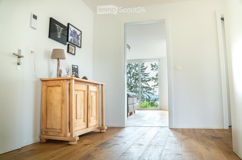 Bright hallway with wooden flooring, white walls, a small wooden console table with a lamp and decorative items, and a doorway leading to a room with a view of greenery.