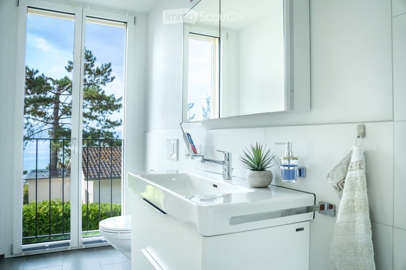 Modern bathroom with white tiles, a large sink, a toilet, a mirror, and a view of the sea through a large window.