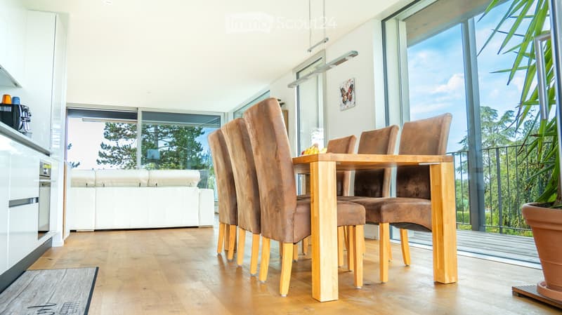 Bright, open-plan living and dining area with wooden flooring, large windows offering a view, a white sofa, a wooden dining table with brown upholstered chairs, and a coffee machine on the counter.