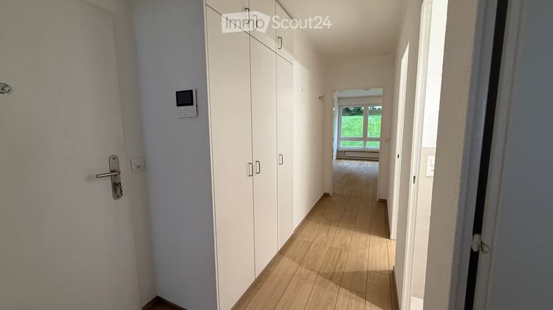 A narrow hallway with light wooden flooring, white walls, built-in white cabinets, and a small digital display mounted on the wall. The hallway leads to a room with large windows and a view of greenery.