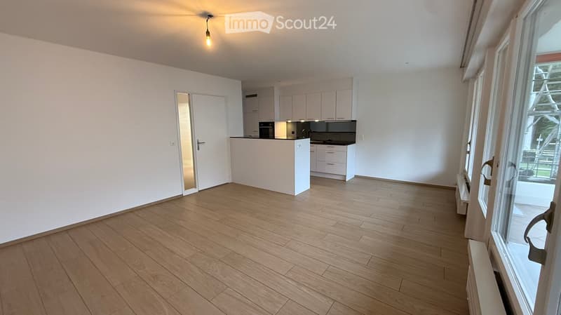 Empty room with wooden flooring, white walls, and a doorway leading to a kitchen area. The kitchen features white cabinets and a countertop.