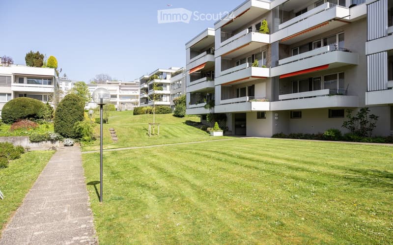 Well-maintained lawn with trimmed hedges, small trees, and a pathway leading to a modern apartment building with balconies and awnings.
