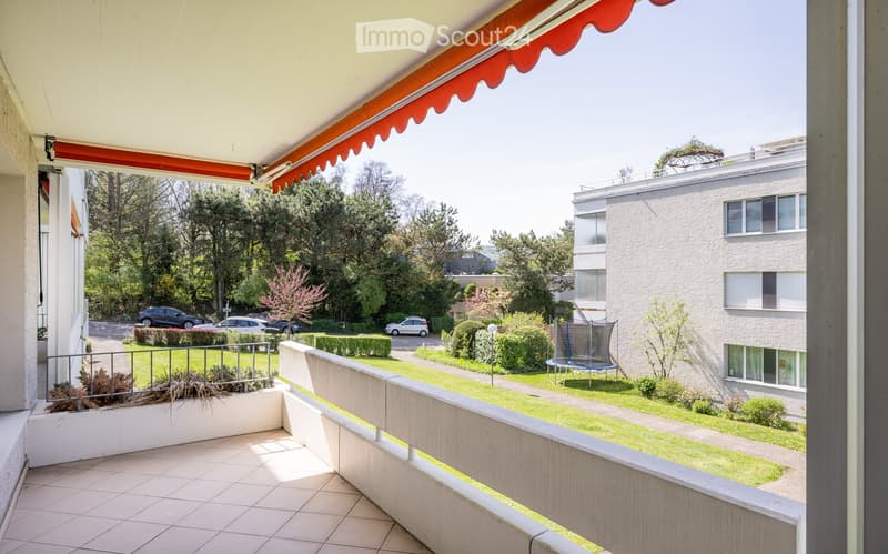 Balcony with tiled floor, metal railing, and an awning. The view shows a landscaped garden area with trees, shrubs, a trampoline, and parked cars.