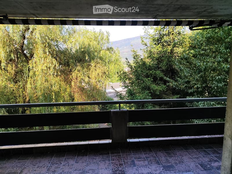Balcony with tiled floor, black metal railing, and lush greenery in the background, including trees and a distant mountain view.