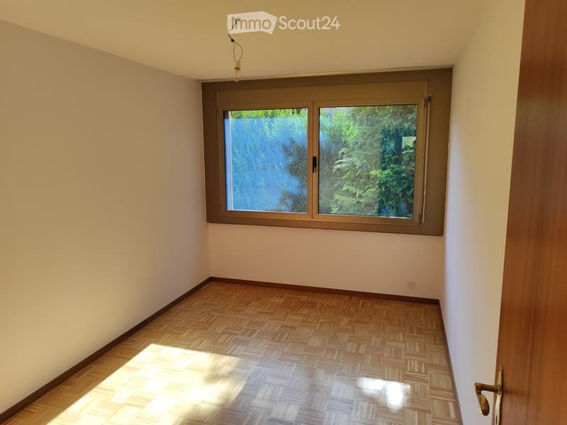 Empty room with wooden parquet flooring, white walls, and a large window with a view of greenery outside.