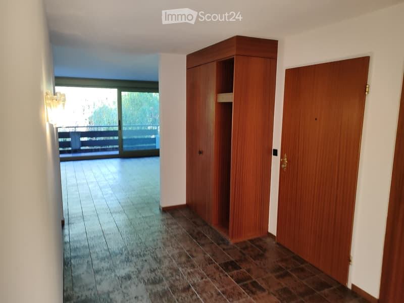 A clean, well-lit hallway with wooden doors, tiled flooring, and a view to a balcony area through large glass doors.