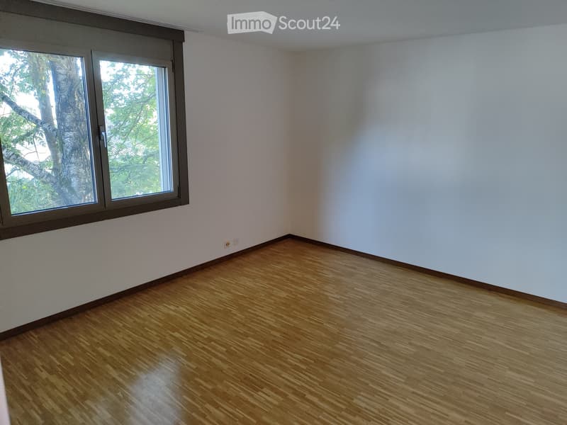 Empty room with wooden flooring and white walls, featuring a large window with a view of trees.
