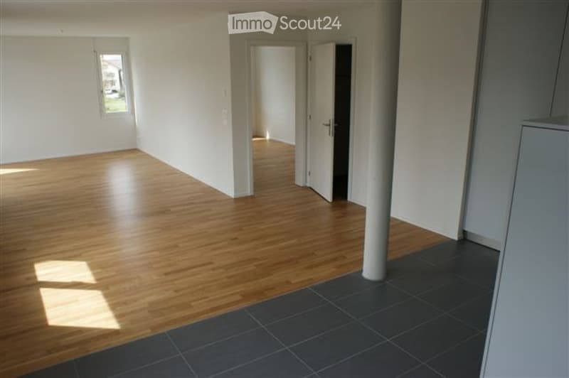 Empty room with wooden flooring, white walls, and a large window allowing natural light. The space features an open doorway leading to another room and a central column.