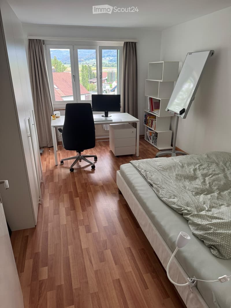A well-lit bedroom with a bed, desk, chair, and bookshelf, featuring large windows with a scenic view.