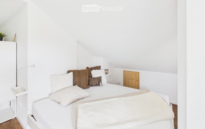A modern, minimalist bedroom with a white bed, multiple pillows, a blanket, a nightstand with a lamp, and a wardrobe.
