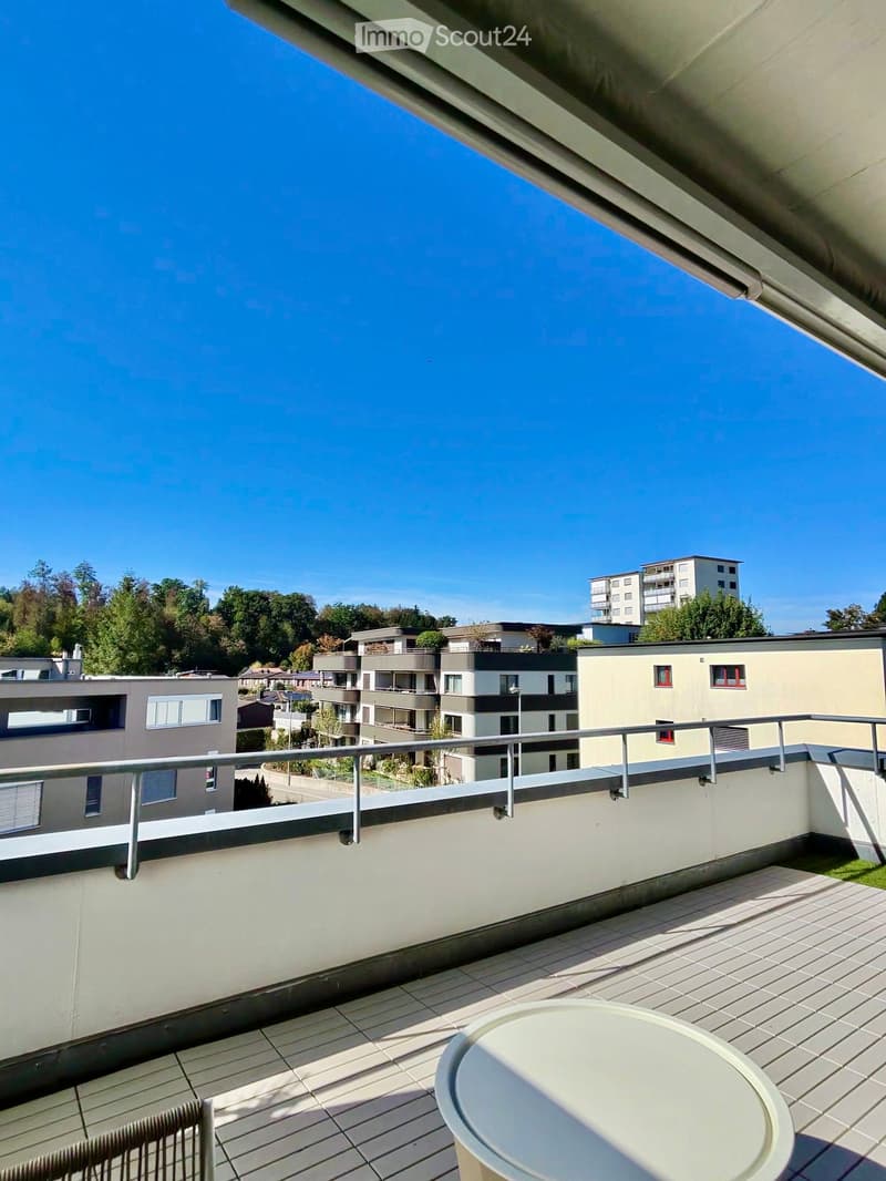 Balcony with tiled floor, metal railing, round table, and chair, overlooking a residential area with modern buildings and trees.