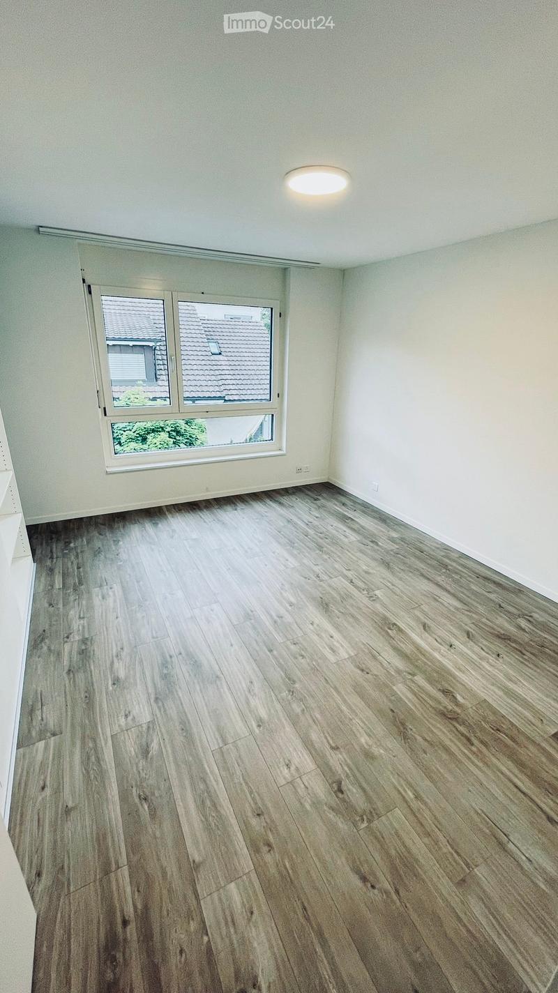 Empty room with light-colored walls, wooden flooring, and a large window with a view of neighboring rooftops.
