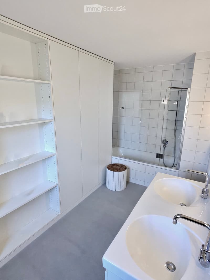 Modern bathroom with white tiles, a double sink, a bathtub, a small round stool, and built-in white cabinets with shelves.