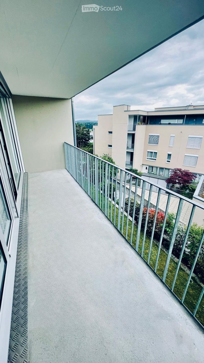 Modern balcony with a metal railing, concrete floor, and a view of neighboring buildings and greenery.
