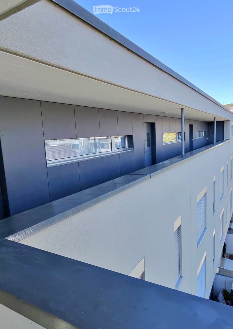 Modern balcony with white walls, large windows, and a sleek metal railing, overlooking a clear blue sky.