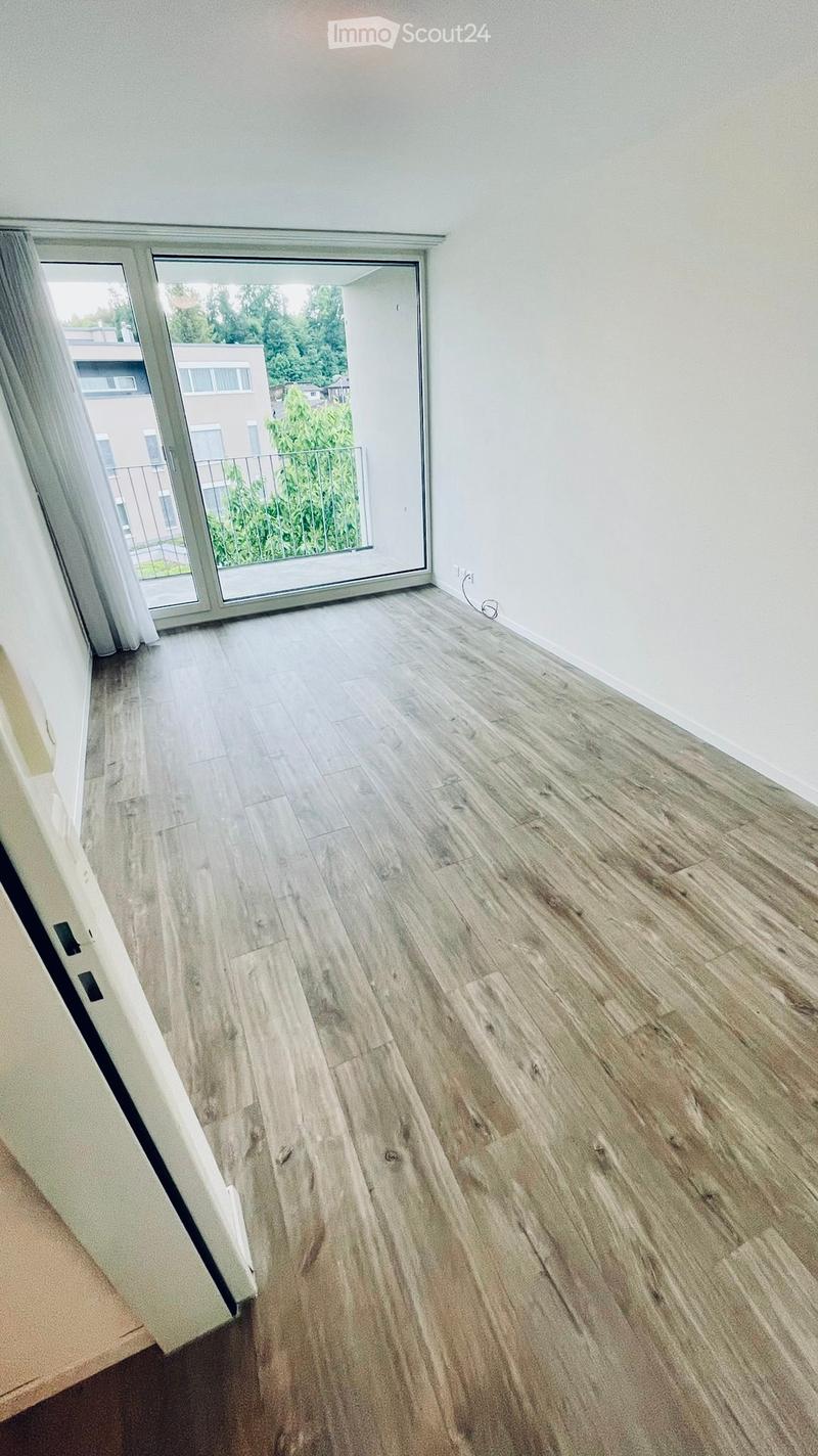 Empty room with light wooden flooring and white walls, featuring a large sliding glass door leading to a balcony with a view of greenery and neighboring buildings.