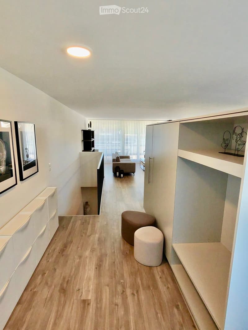 Modern hallway with wooden flooring, white walls, and built-in shelving units. Features a small seating area with a sofa and two round stools, along with decorative mirrors and a shelf with artwork.