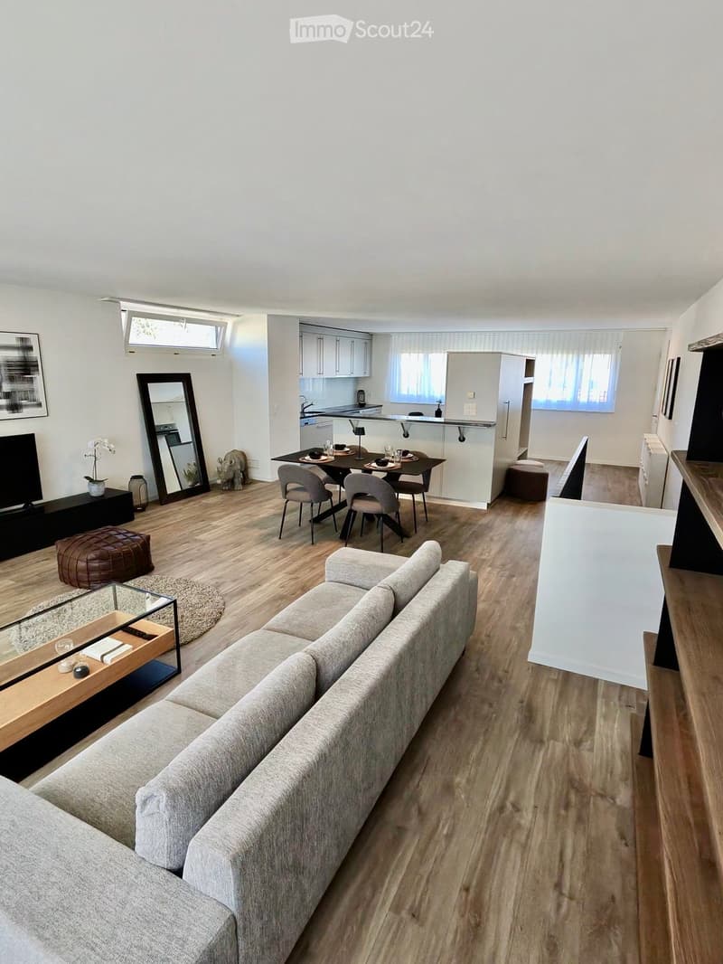Modern living and dining area with a grey sectional sofa, coffee table, ottoman, dining table with chairs, and a small kitchen area with a kitchen island and stools.