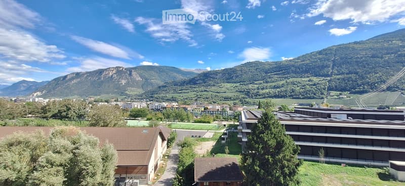 Panoramic view of a town with modern buildings, green spaces, and distant mountains under a clear blue sky.