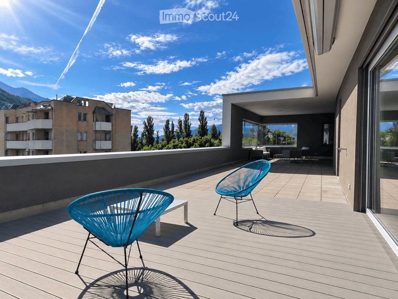 Modern rooftop terrace with wooden decking, two turquoise chairs, and a view of mountains and cityscape.