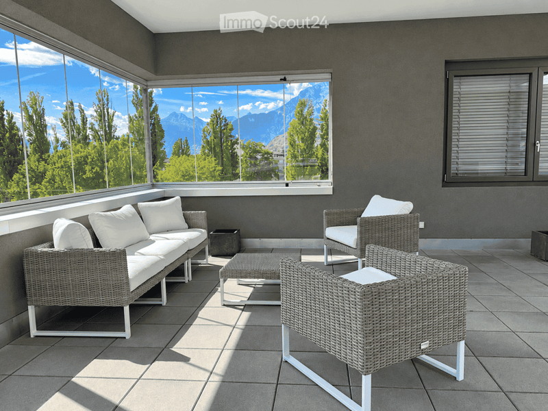 Modern outdoor seating area with wicker furniture, large windows offering a scenic mountain view.