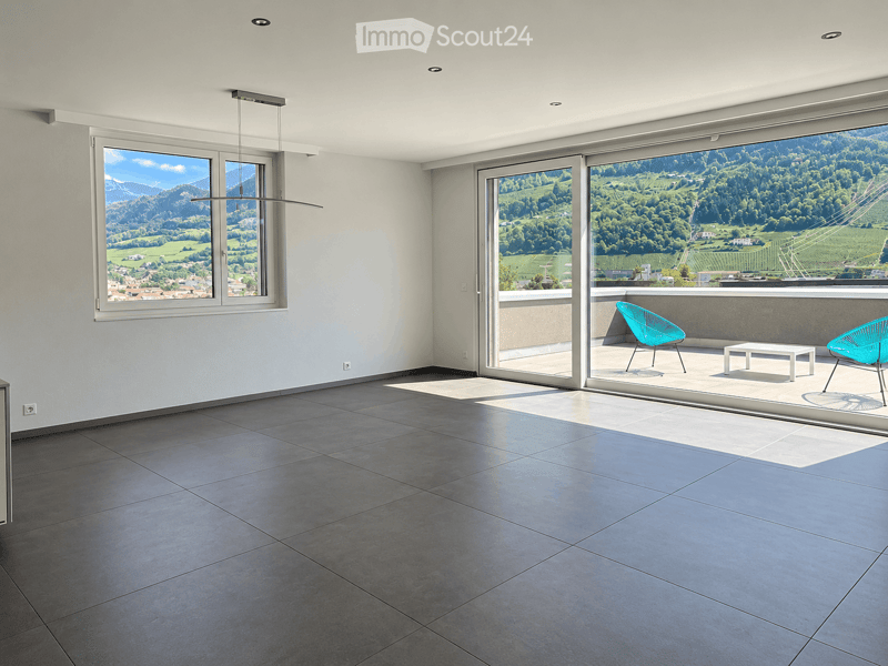 Modern, minimalist room with large windows and sliding glass doors leading to a balcony. The balcony features two turquoise chairs and a small white table, overlooking a scenic valley view.